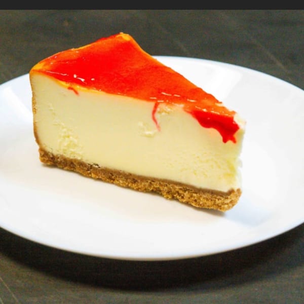 A slice of cheesecake with a graham cracker crust and bright red strawberry topping on a white plate.